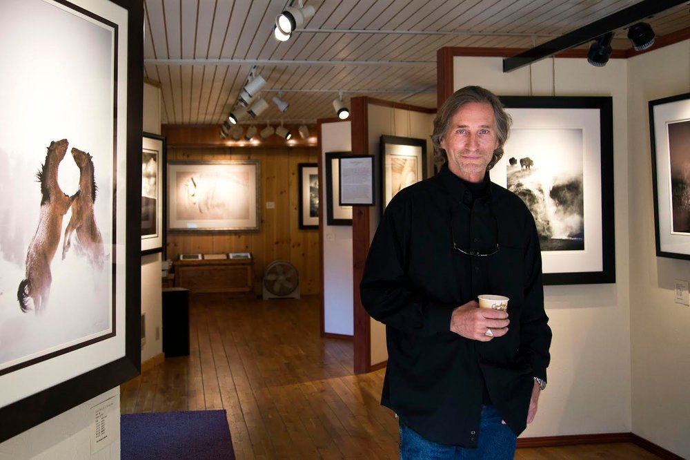 A man holding a cup of coffee stops to look at the camera in an art gallery filled with photographic works