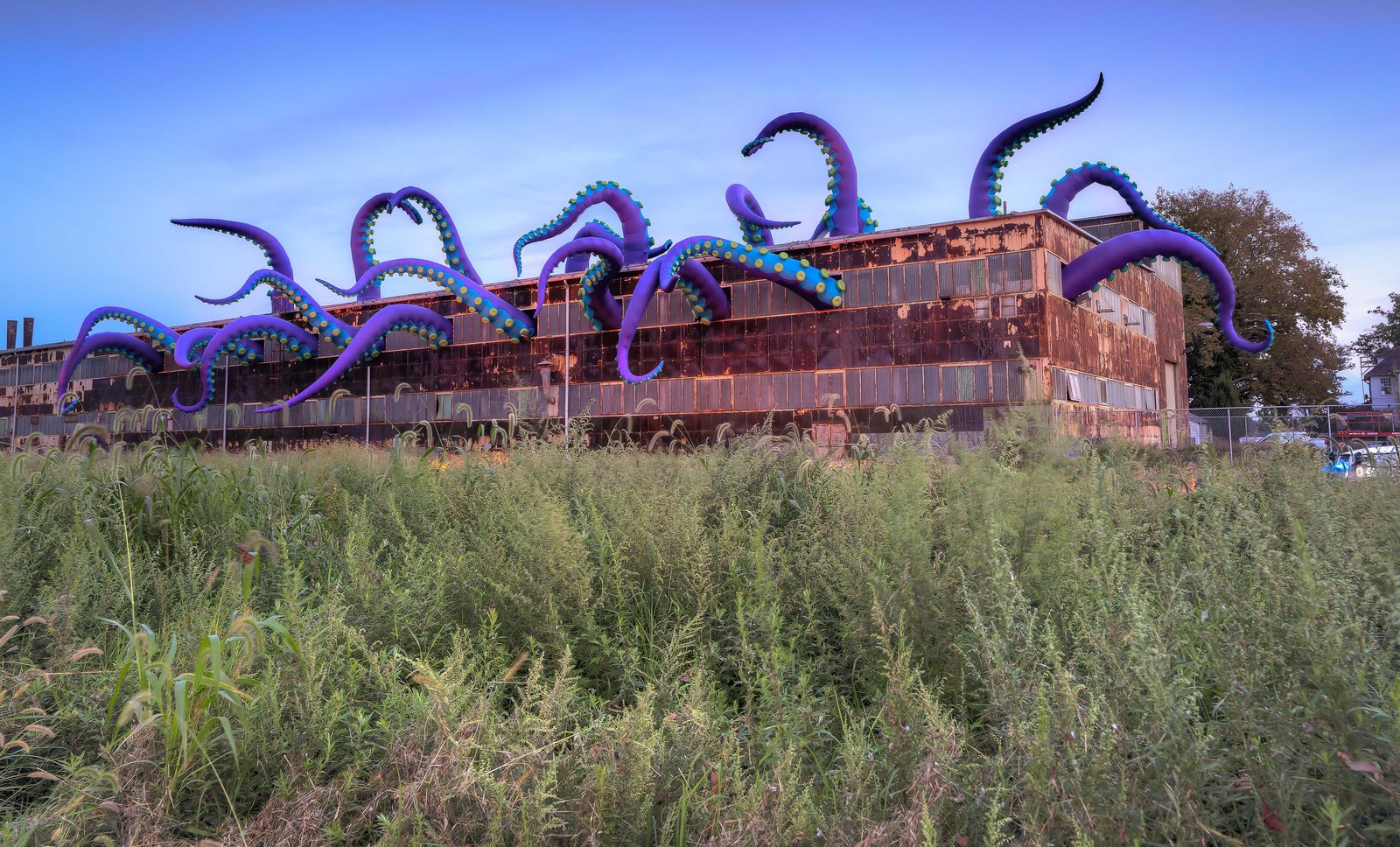 Giant purple and green tentacles swarm out what looks to be an abandoned factory ina grassy field