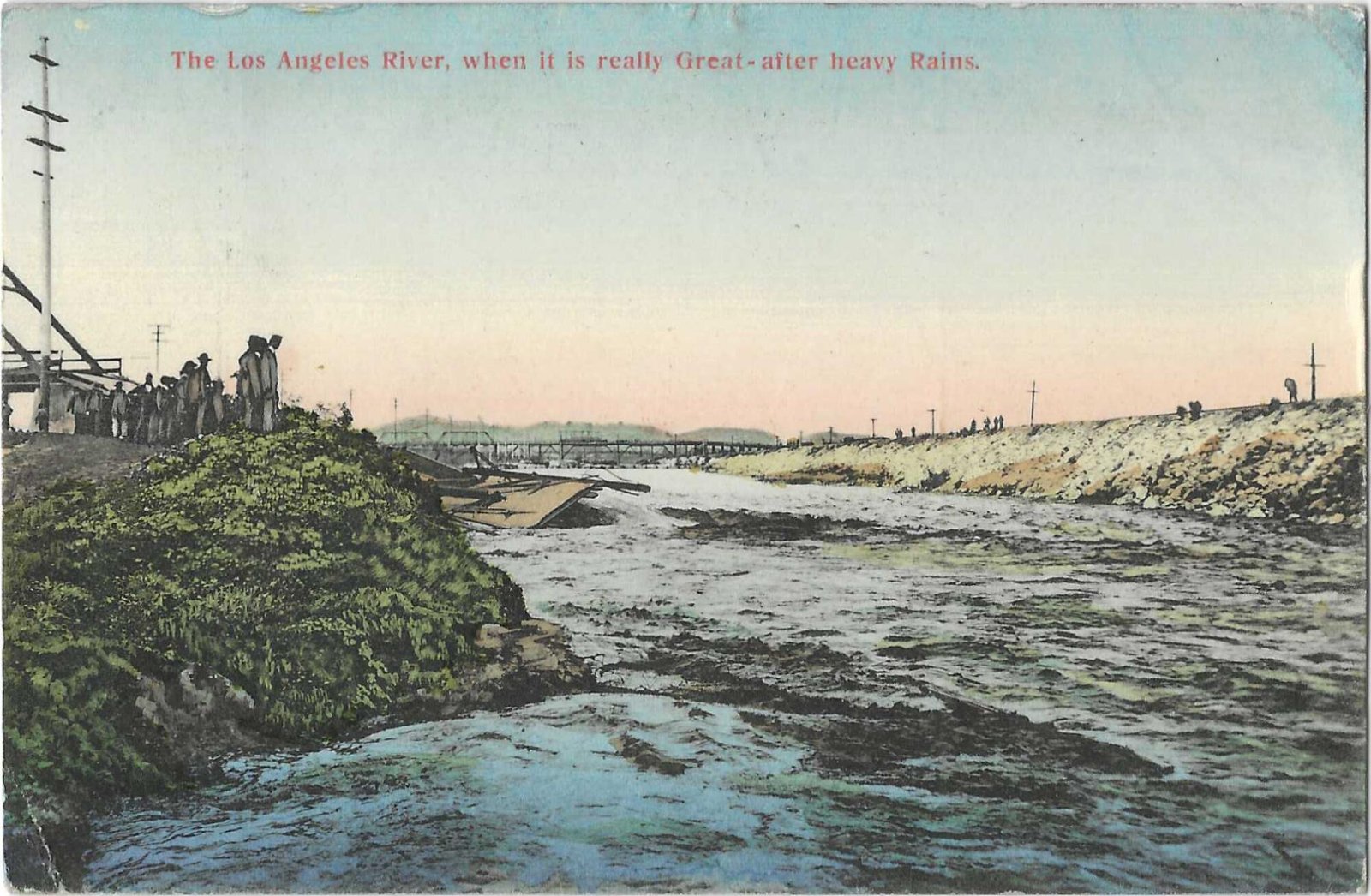Water rushes in the L.A. River basin, with a bridge in the background and people on either bank.