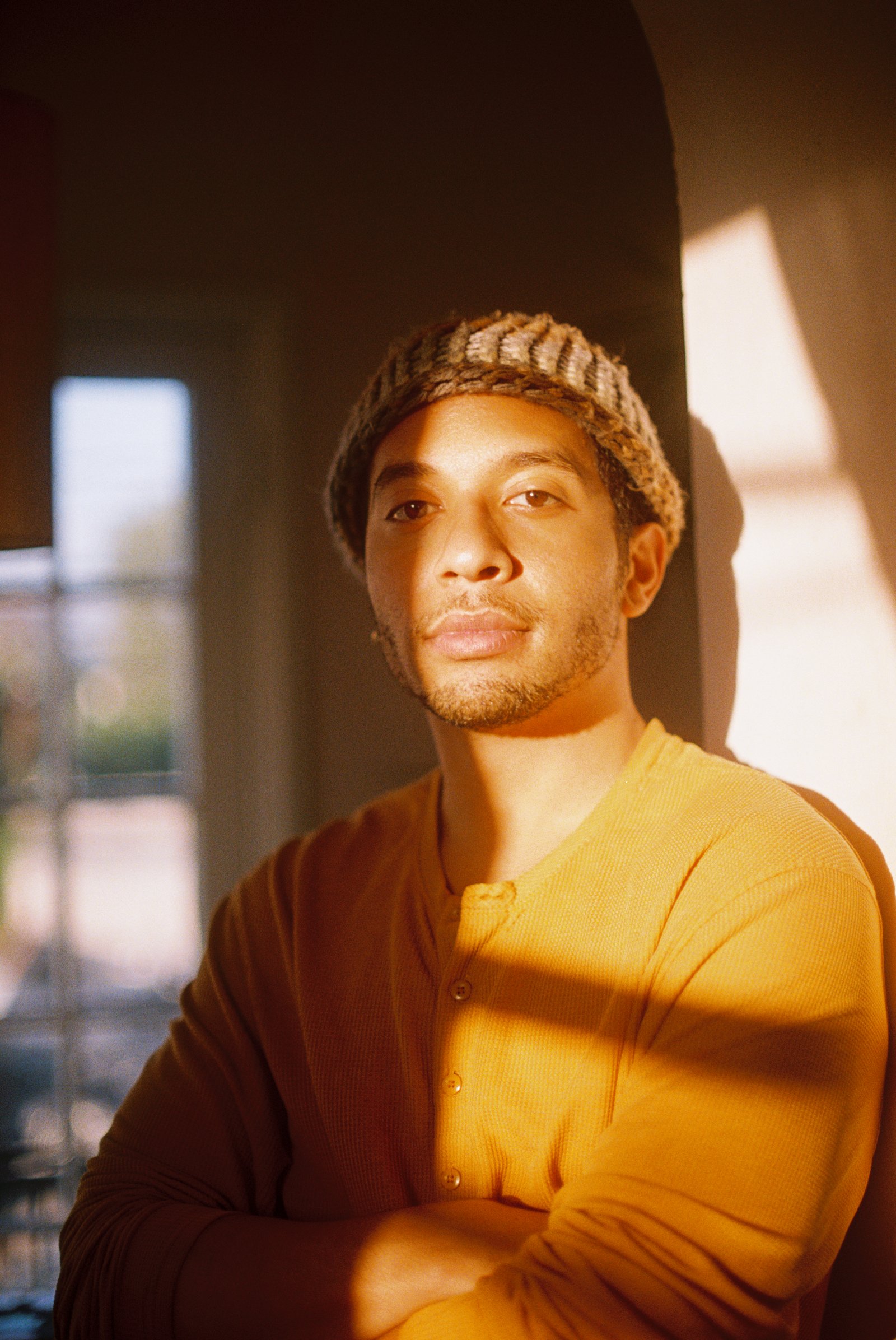 A man wearing a knit beanie and a sweater stands half in and half out of golden light.