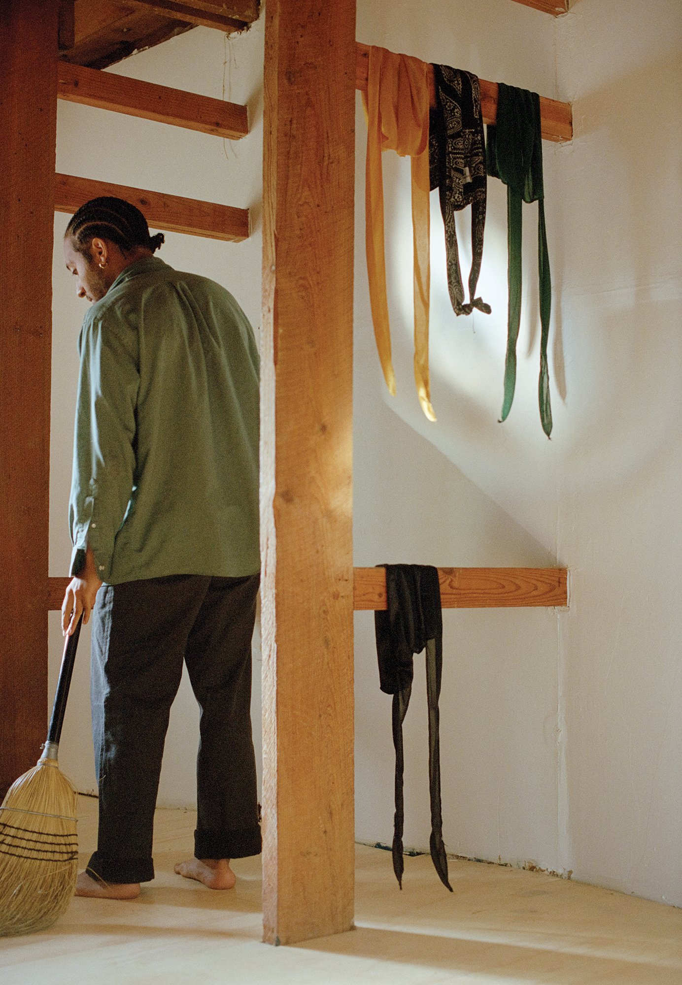 A photograph of a man wearing an army green shirt and dark pants sweeping with his back facing the viewer in a room where several items of clothing hang like ribbons from wooden beams.